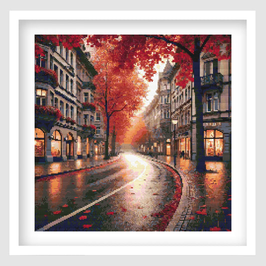 Autumn city street cross stitch pattern featuring a glowing rainy road lined with buildings, shopfronts and red-orange trees, with warm reflections on the pavement.