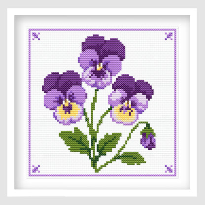 Cross-stitch-style artwork featuring three purple and lavender pansy flowers with yellow centers, green stems and leaves, and a small unopened flower bud. The bouquet is arranged on a white stitched background with a delicate purple border and decorative corner motifs.