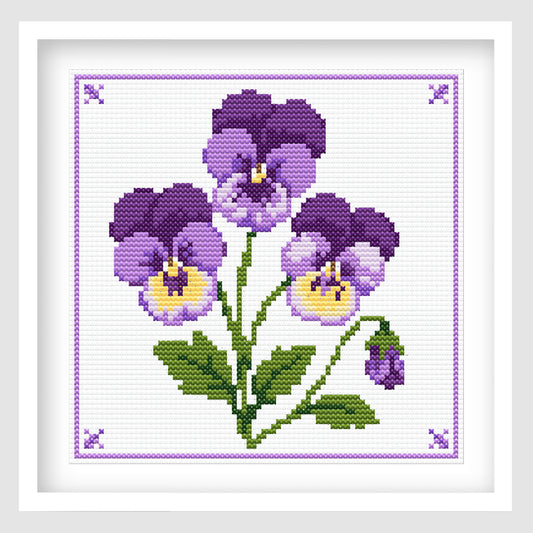 Cross-stitch-style artwork featuring three purple and lavender pansy flowers with yellow centers, green stems and leaves, and a small unopened flower bud. The bouquet is arranged on a white stitched background with a delicate purple border and decorative corner motifs.