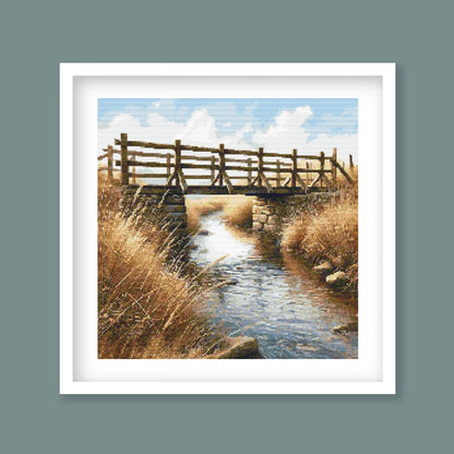 Framed artwork of a wooden bridge over a stream with tall grass and blue sky.