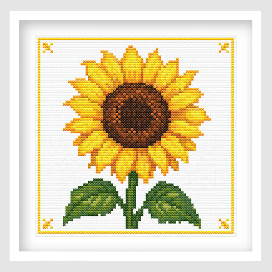 Cross-stitch-style artwork of a bright yellow sunflower with a large textured brown center, surrounded by green leaves and a central stem. The flower is centered on a white stitched background with a delicate yellow border and decorative corner motifs.
