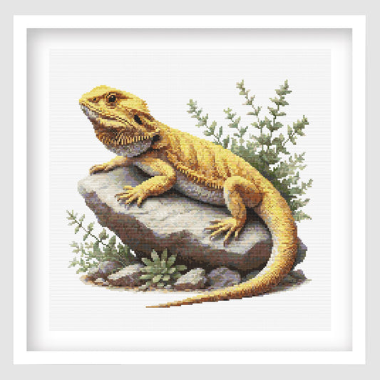 Cross-stitched yellow bearded dragon lizard on a rock with green plants.
