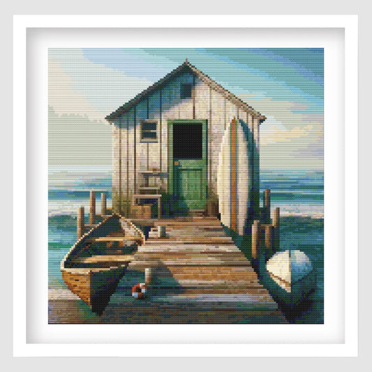 Boat Shed Cross Stitch Pattern displayed as framed wall art with a weathered coastal shed, wooden jetty, rowboat, and seaside detail.