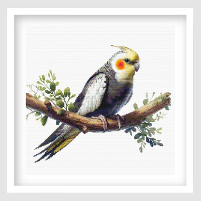 Cockatiel cross stitch pattern featuring a grey and yellow cockatiel with an orange cheek patch perched on a leafy branch, shown as framed wall art.