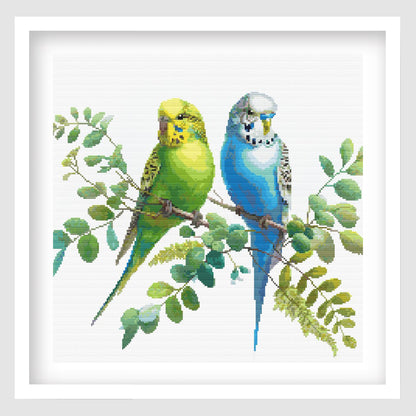 Colorful Budgies Cross Stitch Pattern displayed as framed wall art with two bright budgies perched among soft green foliage.