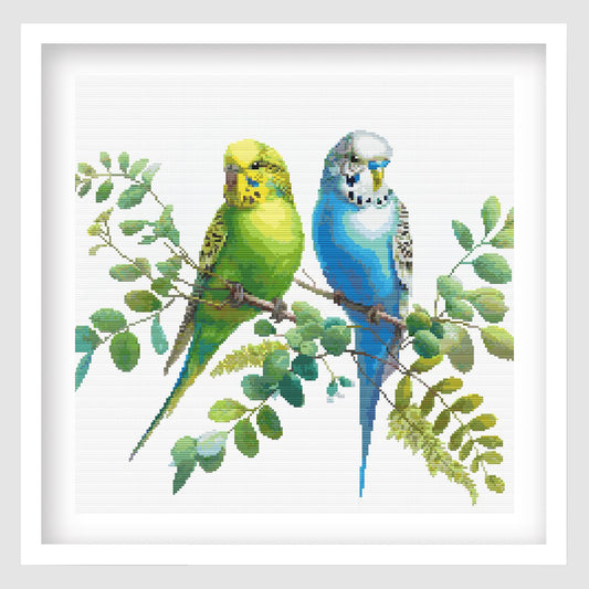 Colorful Budgies Cross Stitch Pattern displayed as framed wall art with two bright budgies perched among soft green foliage.