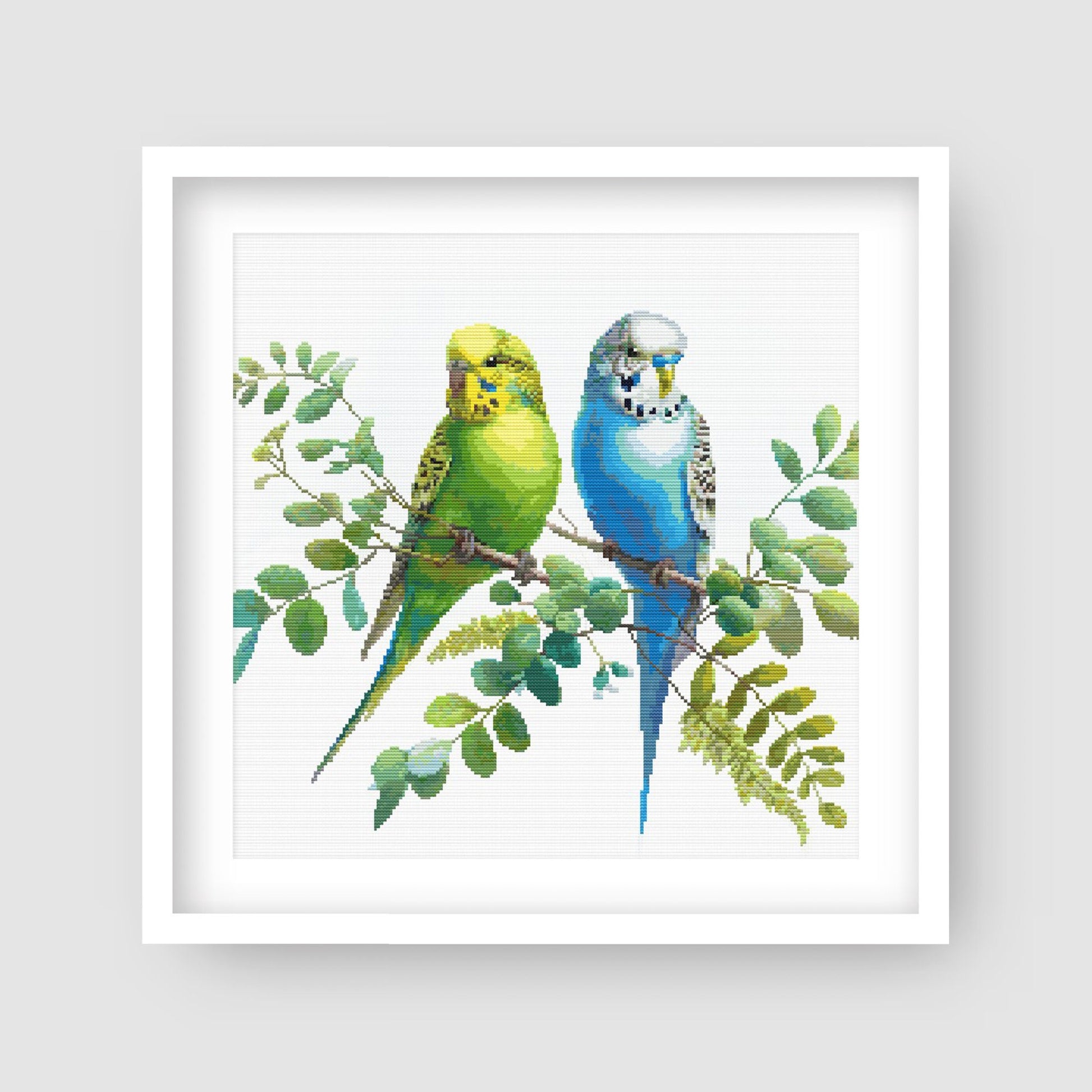 Colorful Budgies Cross Stitch Pattern displayed in a white frame as pet bird wall art with two colourful budgies and soft foliage.