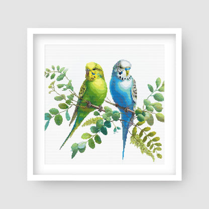 Colorful Budgies Cross Stitch Pattern displayed in a white frame as pet bird wall art with two colourful budgies and soft foliage.