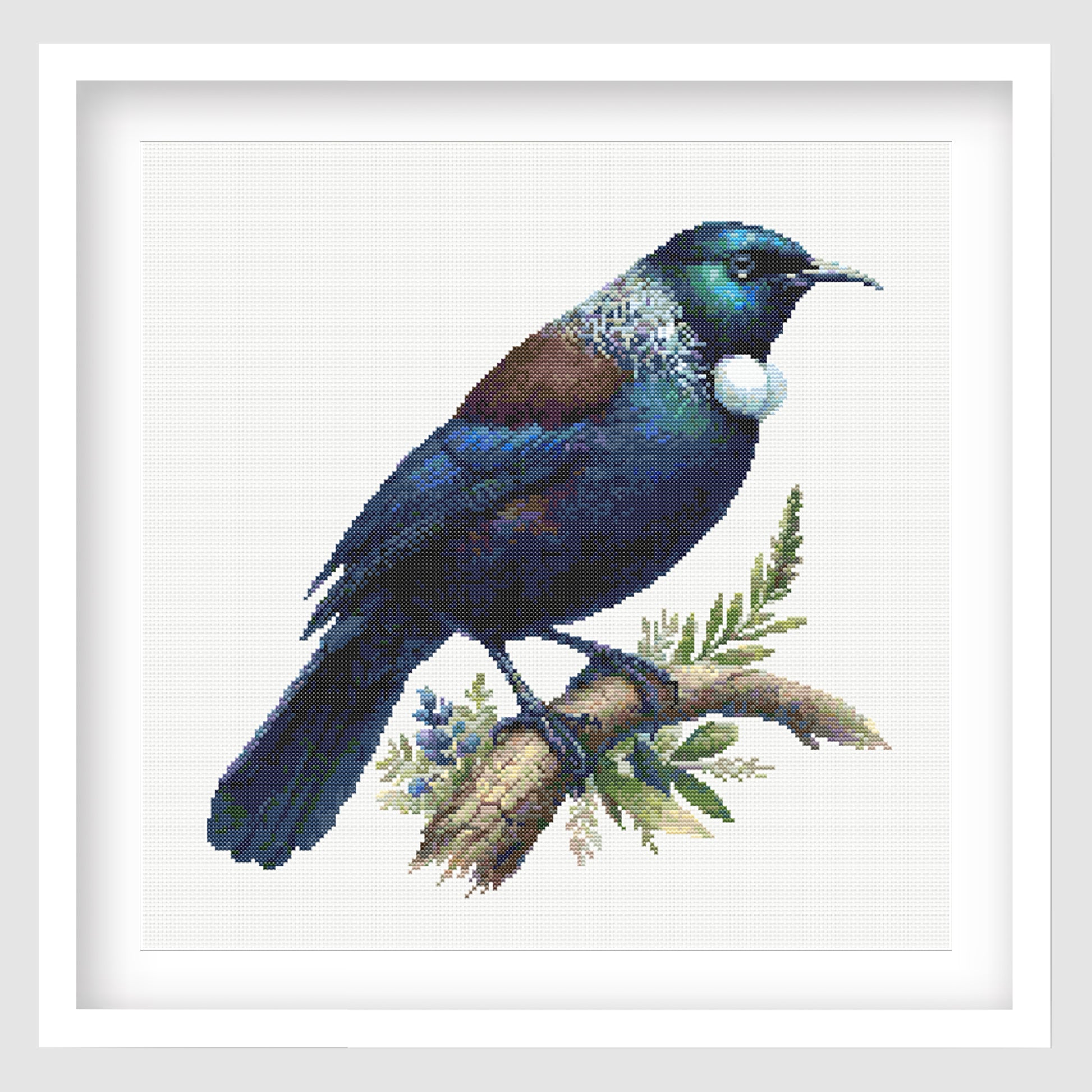 New Zealand Tui Cross Stitch Pattern displayed as framed wall art with a native tui bird, glossy blue-green feathers, white throat tuft, and natural branch detail.Colorful bird illustration on a branch with leaves, framed by a white border.