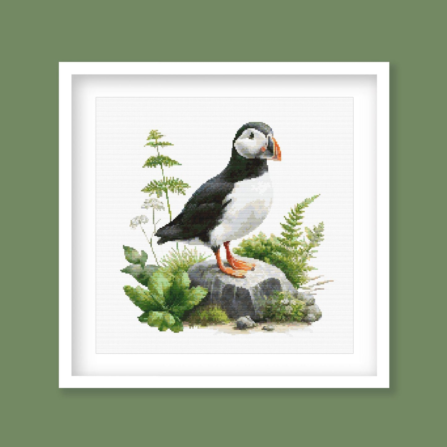 Puffin Cross Stitch Pattern displayed in a green frame as coastal bird wall art with natural foliage and puffin detail.