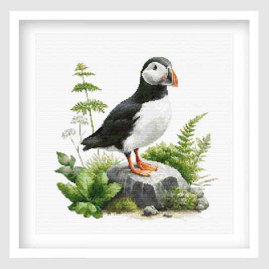 Puffin Cross Stitch Pattern displayed as framed wall art with a coastal puffin, bright orange beak, green foliage, rocks, and natural bird detail.