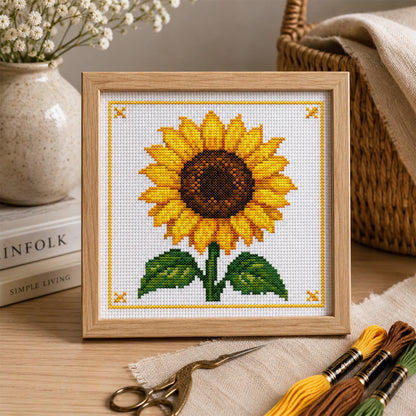 Sunflower cross stitch pattern featuring a bright yellow sunflower with green leaves and a decorative yellow stitched border, displayed as framed wall art.